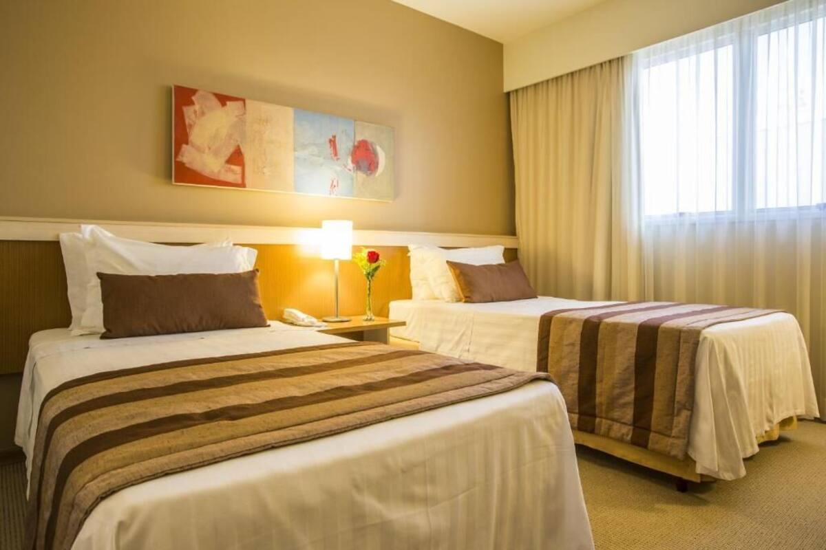 Quarto de hotel com duas camas, uma mesa de cabeceira com abajur, um vaso com flor e um quadro na parede.