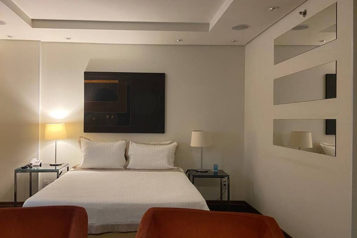 Quarto de hotel com uma cama grande, duas mesas de cabeceira, um espelho fixado na parede e duas cadeiras.