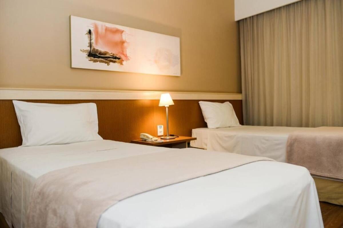 Quarto de hotel com duas camas, uma mesa de cabeceira com abajur e um quadro na parede.