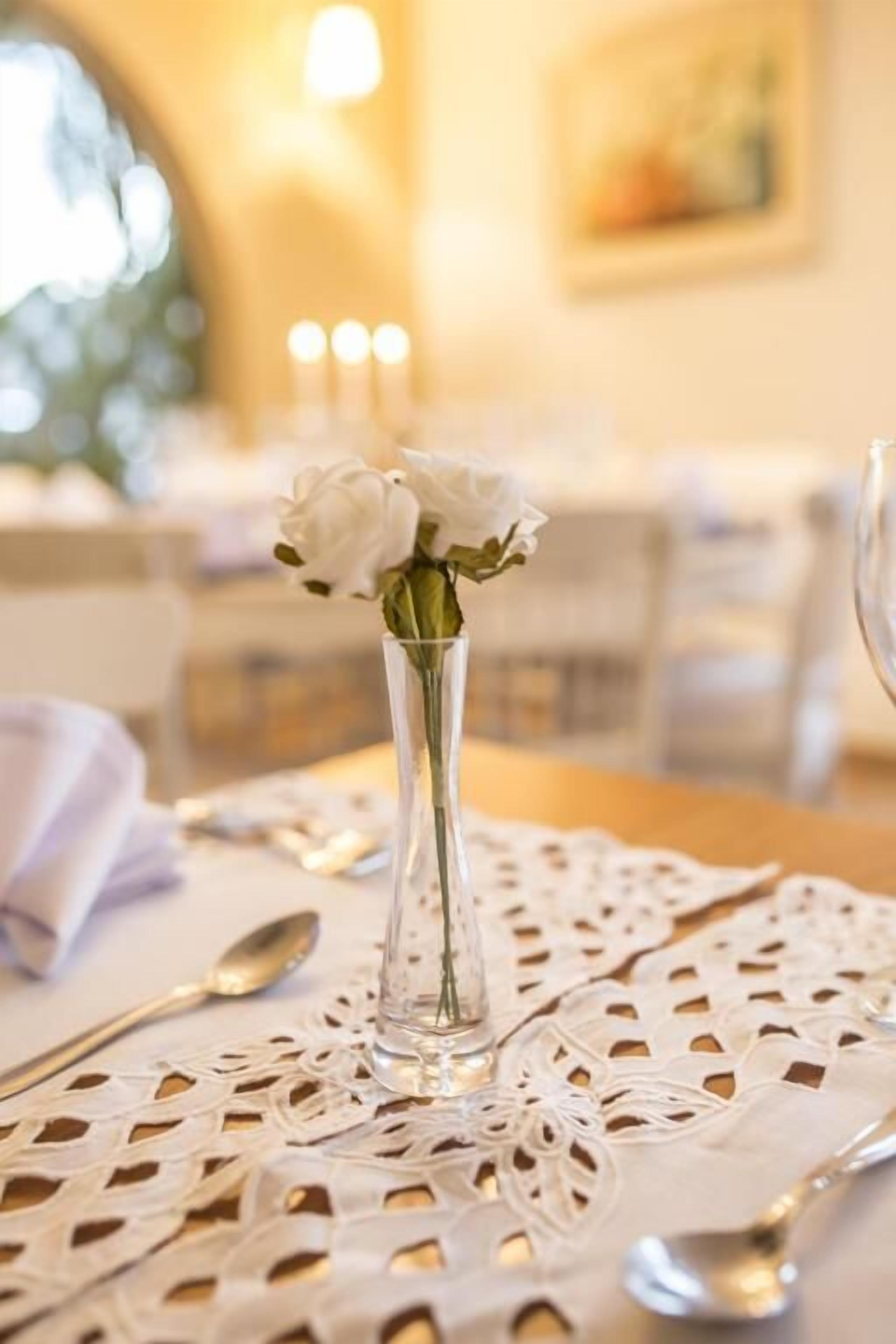 A dining table with a lace tablecloth, a clear glass vase with white flowers, and a spoon.