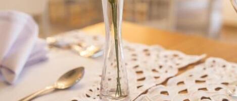 A dining table with a lace tablecloth, a clear glass vase with white flowers, and a spoon.