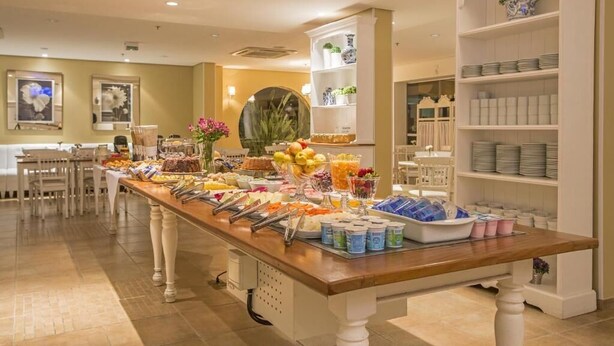 A buffet setup with various food items and beverages, a white shelving unit with decorative items, and a dining area with chairs and tables. - Astron Hotel Bauru by Nobile (Bauru)