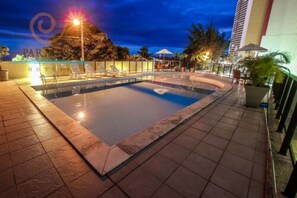 A swimming pool with lounge chairs and umbrellas, surrounded by a tiled deck and a building in the background. - Paradise Flat (Natal)