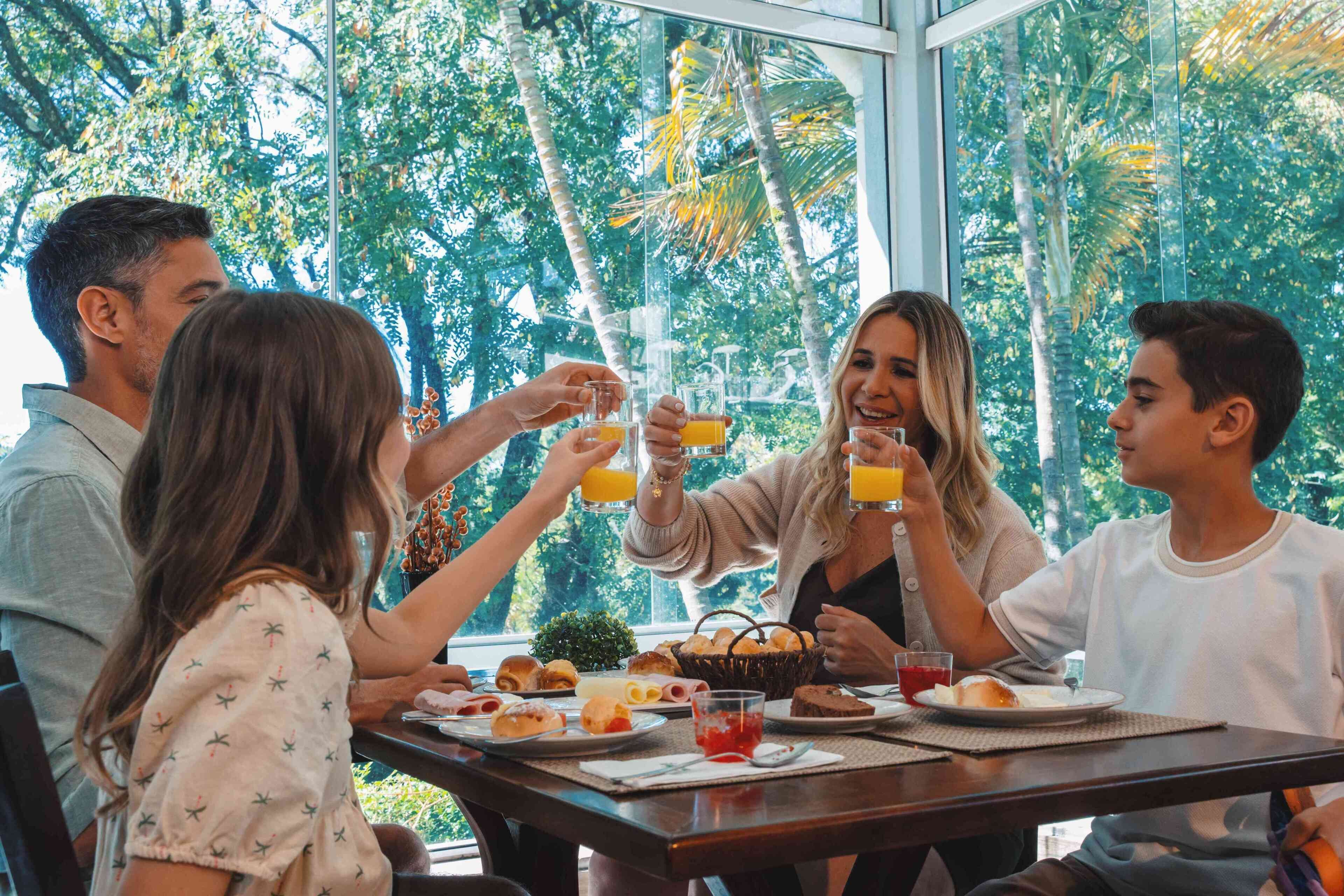 A group of people toasting with orange juice at a table with breakfast items, in a room with large windows and a view of trees.