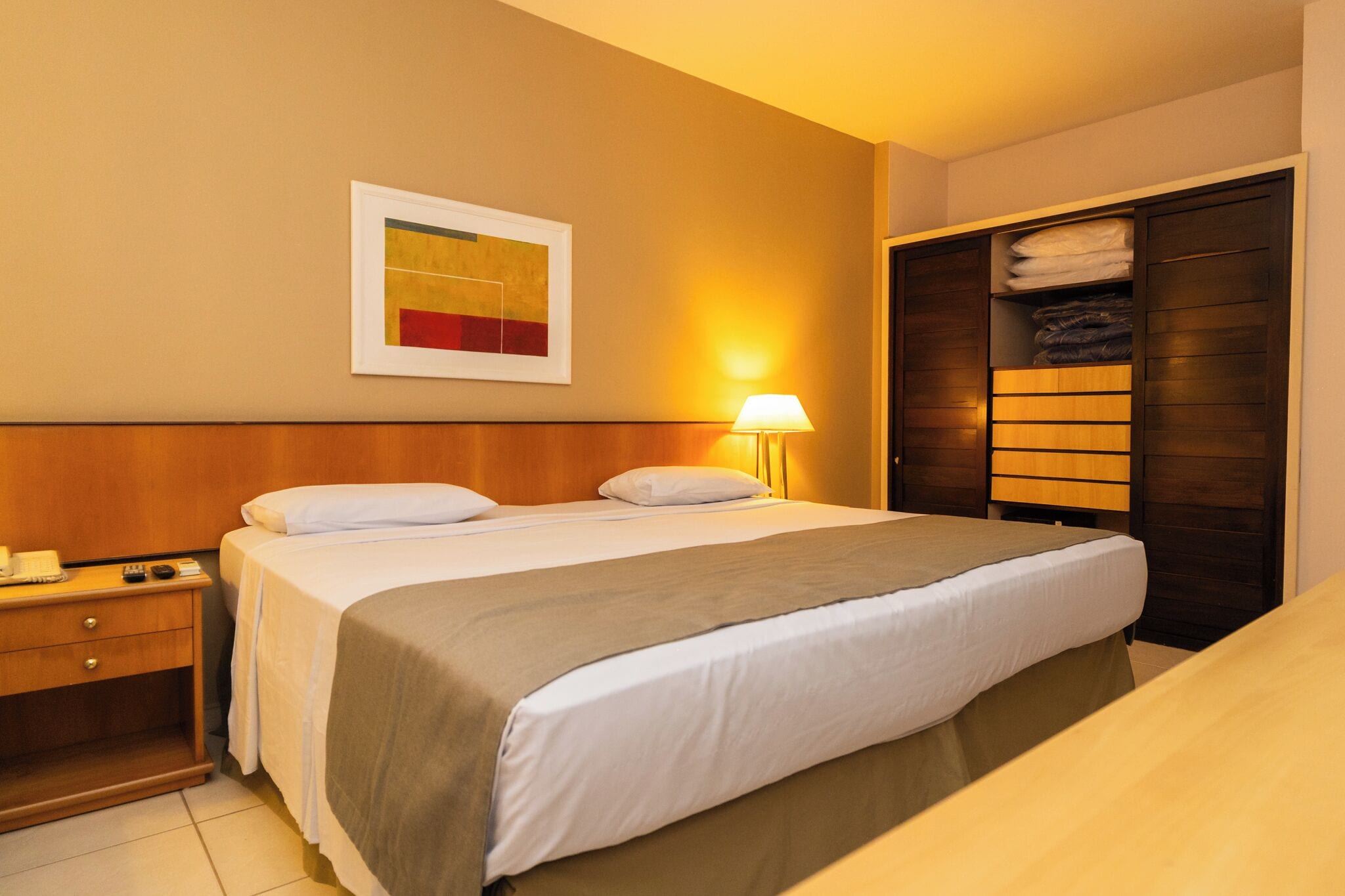 Quarto de hotel com cama, mesa de cabeceira, abajur e guarda-roupa.