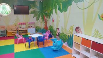 Uma sala de jogos infantil com tapetes de espuma coloridos, uma mesa redonda, cadeiras, uma cozinha de brinquedo e um mural com tema de selva.
