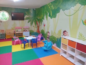 Un cuarto de juegos infantil con alfombras de espuma de colores, una mesa redonda, sillas, una cocina de juguete y un mural con una escena de selva.