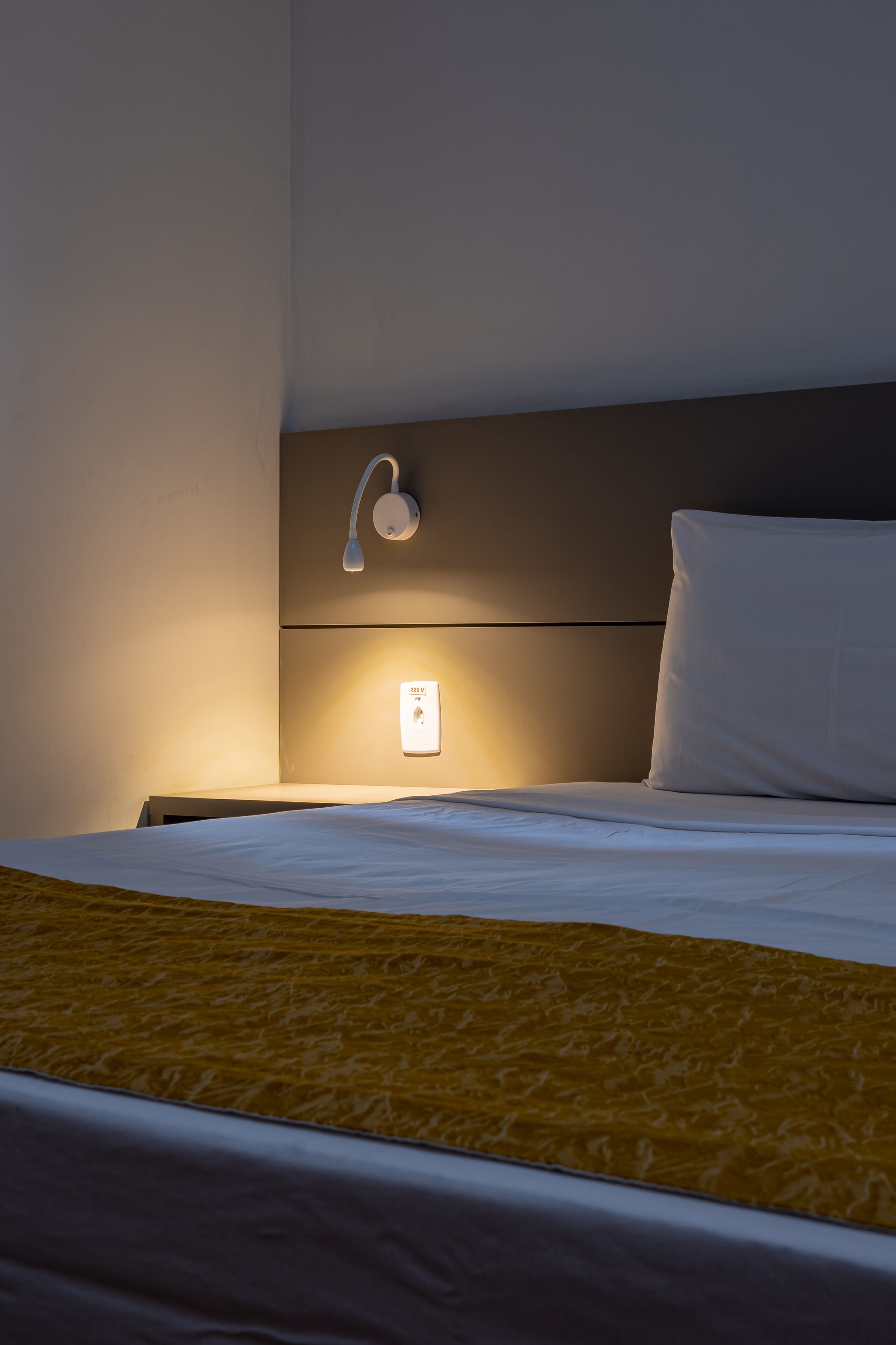 A neatly made bed with a white pillow and a yellow blanket, a wall-mounted light, and a power outlet on the wall.