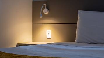 A neatly made bed with a white pillow and a yellow blanket, a wall-mounted light, and a power outlet on the wall.
