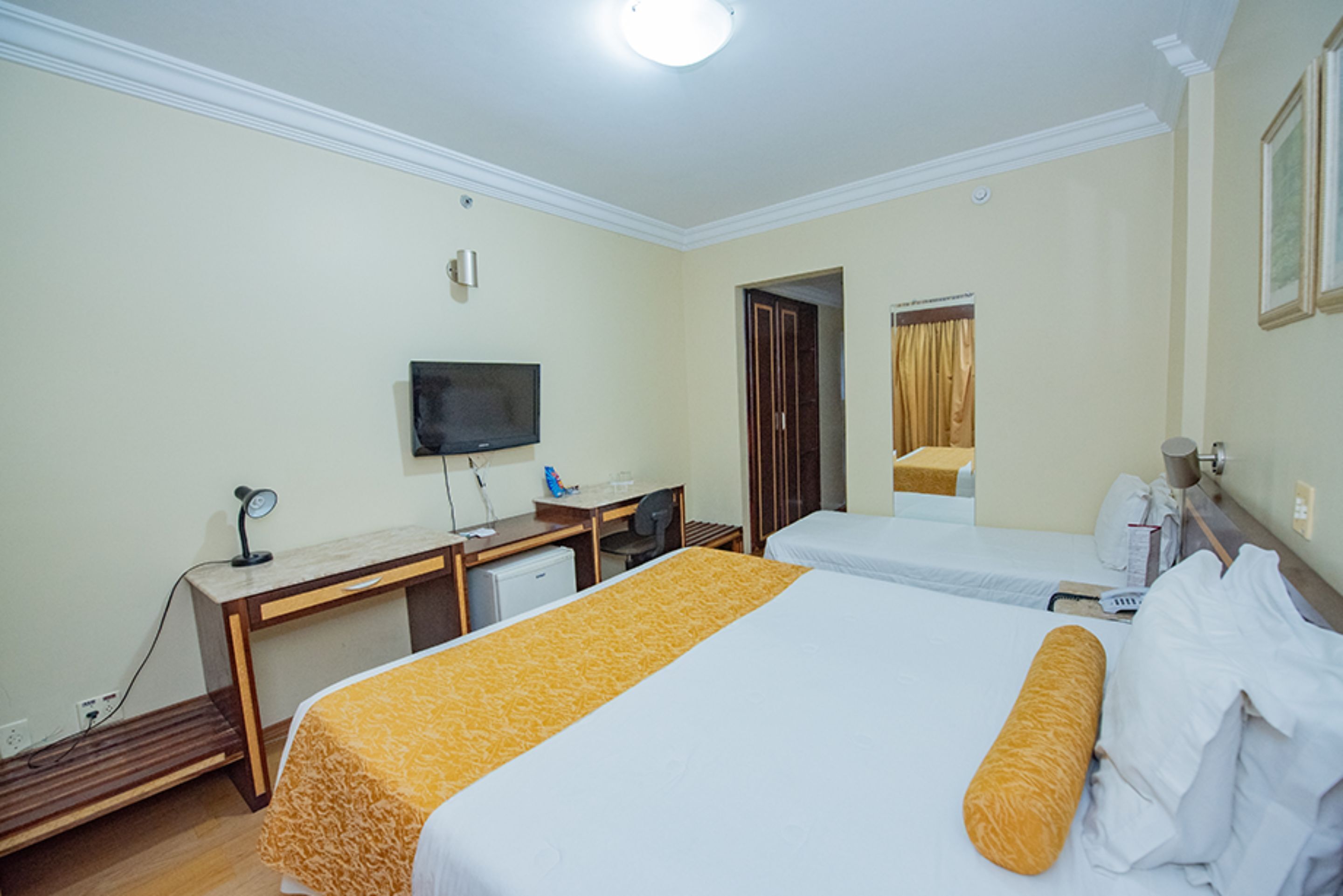 A hotel room with two beds, a desk, a television, and a small kitchenette.