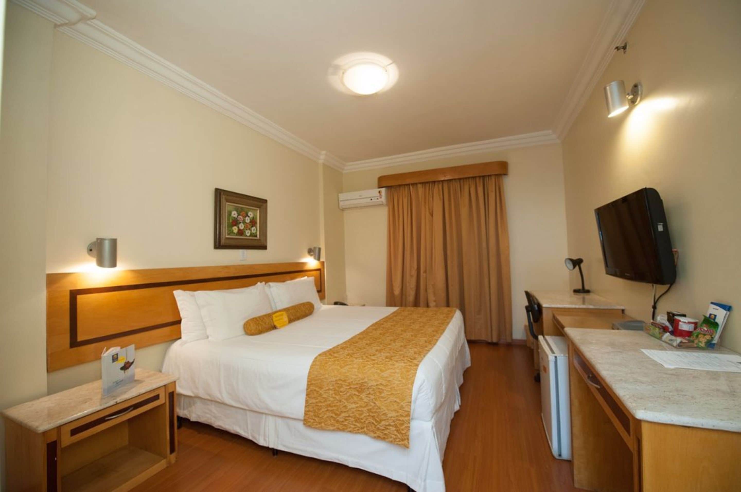 A hotel room with a bed, bedside table, desk, chair, television, and a window with curtains.