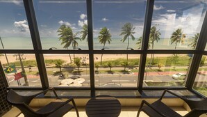 Un balcón con sillas de mimbre y una pequeña mesa, con vista a una playa con palmeras y un cielo despejado.