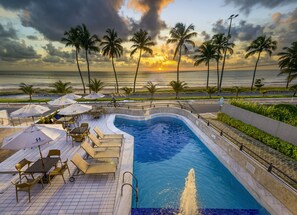 A pool area with lounge chairs and umbrellas, a fountain, and a view of the ocean and palm trees.