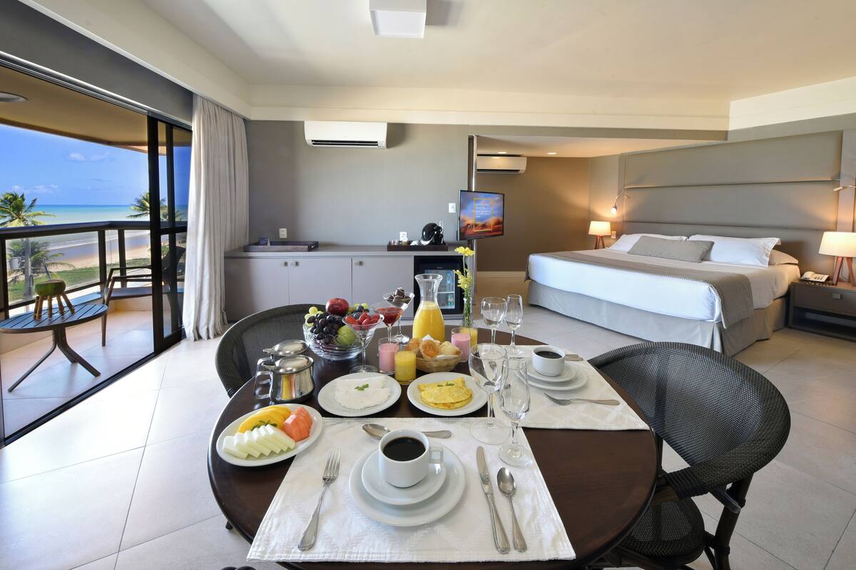 Habitación de hotel con balcón, zona de comedor con desayuno, cama y televisor.
