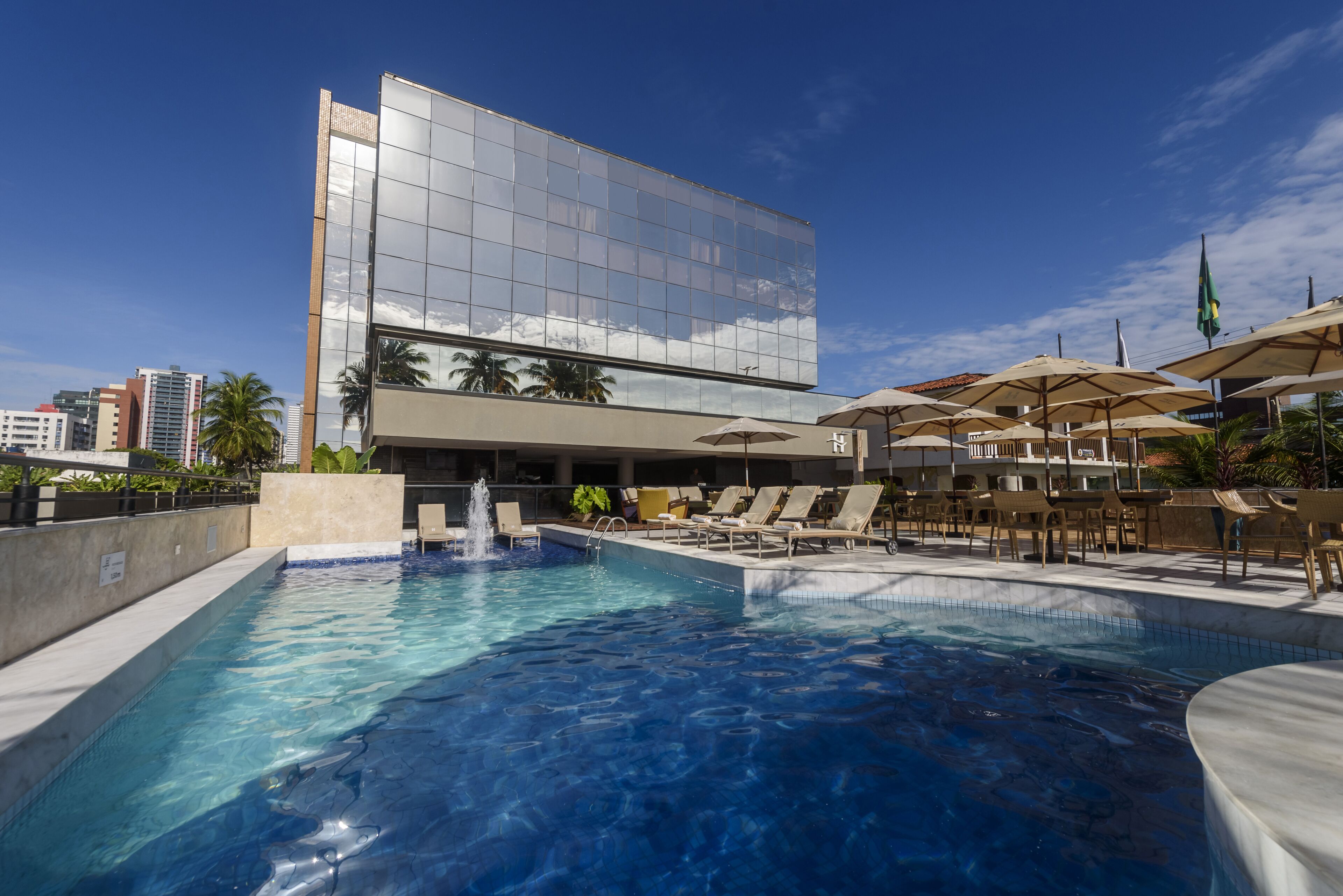 A modern hotel with a large reflective glass building, a swimming pool, and outdoor seating with umbrellas.