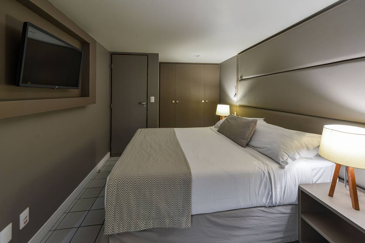 Una habitación de hotel con una cama, televisión fijada en la pared, lámpara de noche y armario.