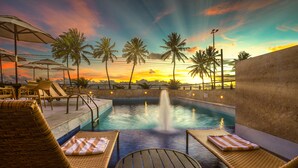 Un área de piscina con sillones de descanso y sombrillas, una fuente y vistas al atardecer.
