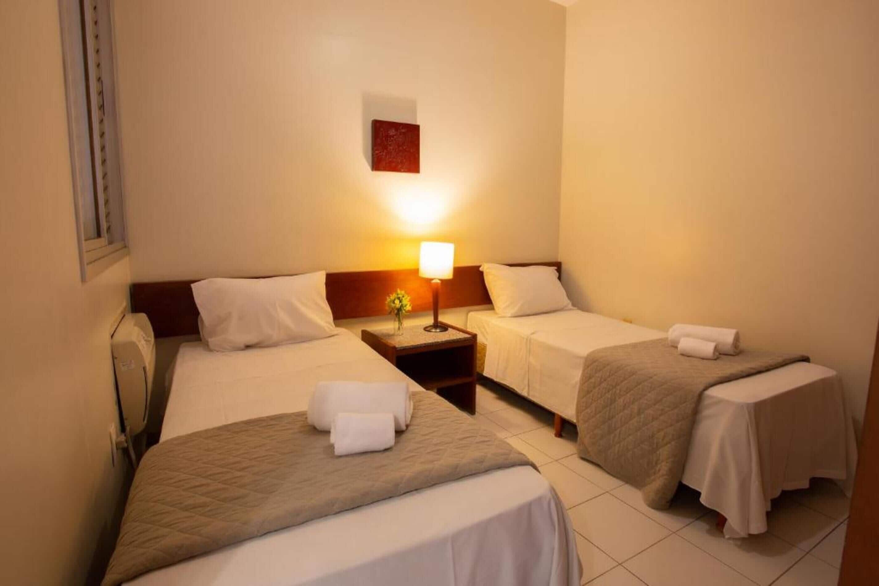 Habitación de hotel con dos camas individuales, una mesita de noche con lámpara y una pequeña mesa con un florero.