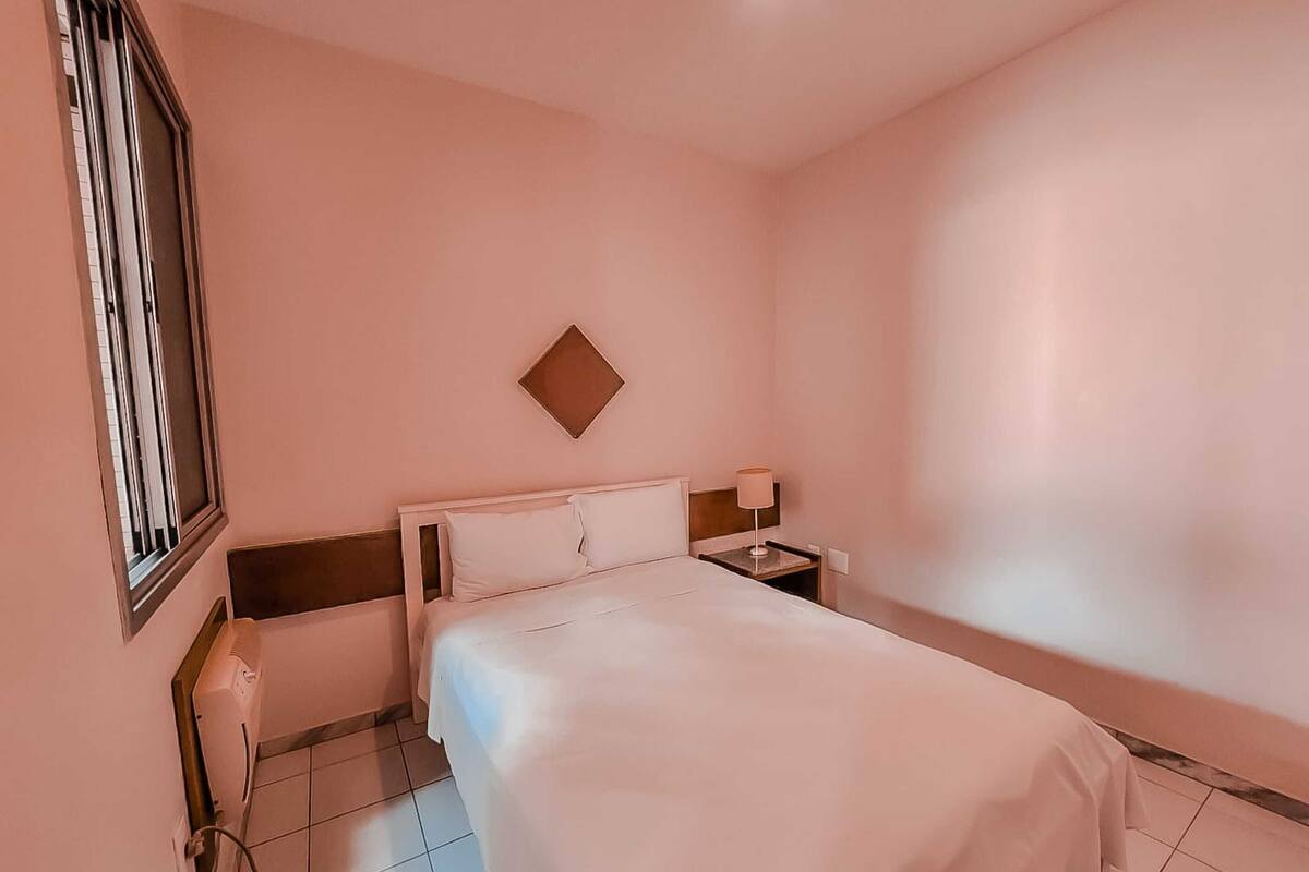 Una camera d'albergo con un letto, un comodino, una lampada, una TV a parete e una finestra con persiane.