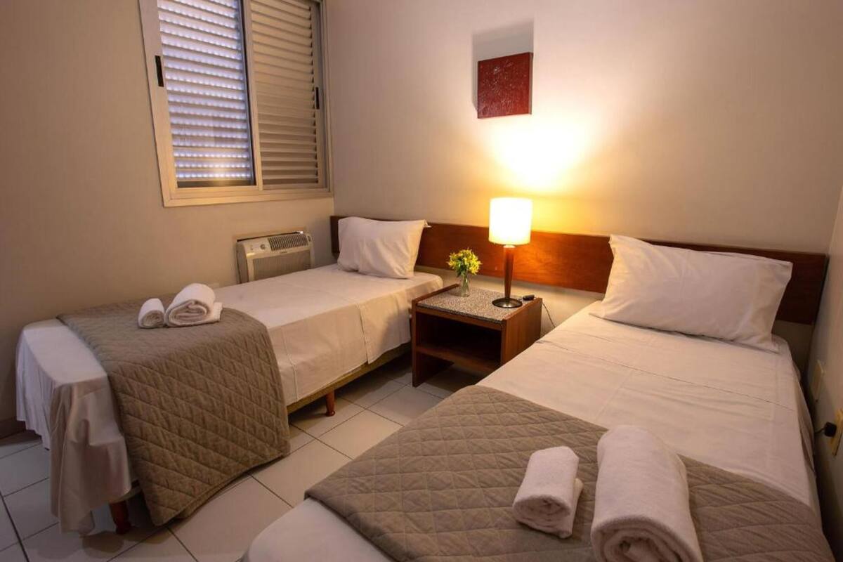 Una camera d'albergo con due letti, ognuno con copripiumino trapuntato e un asciugamano arrotolato sopra. Tra i letti, un comodino con una lampada.