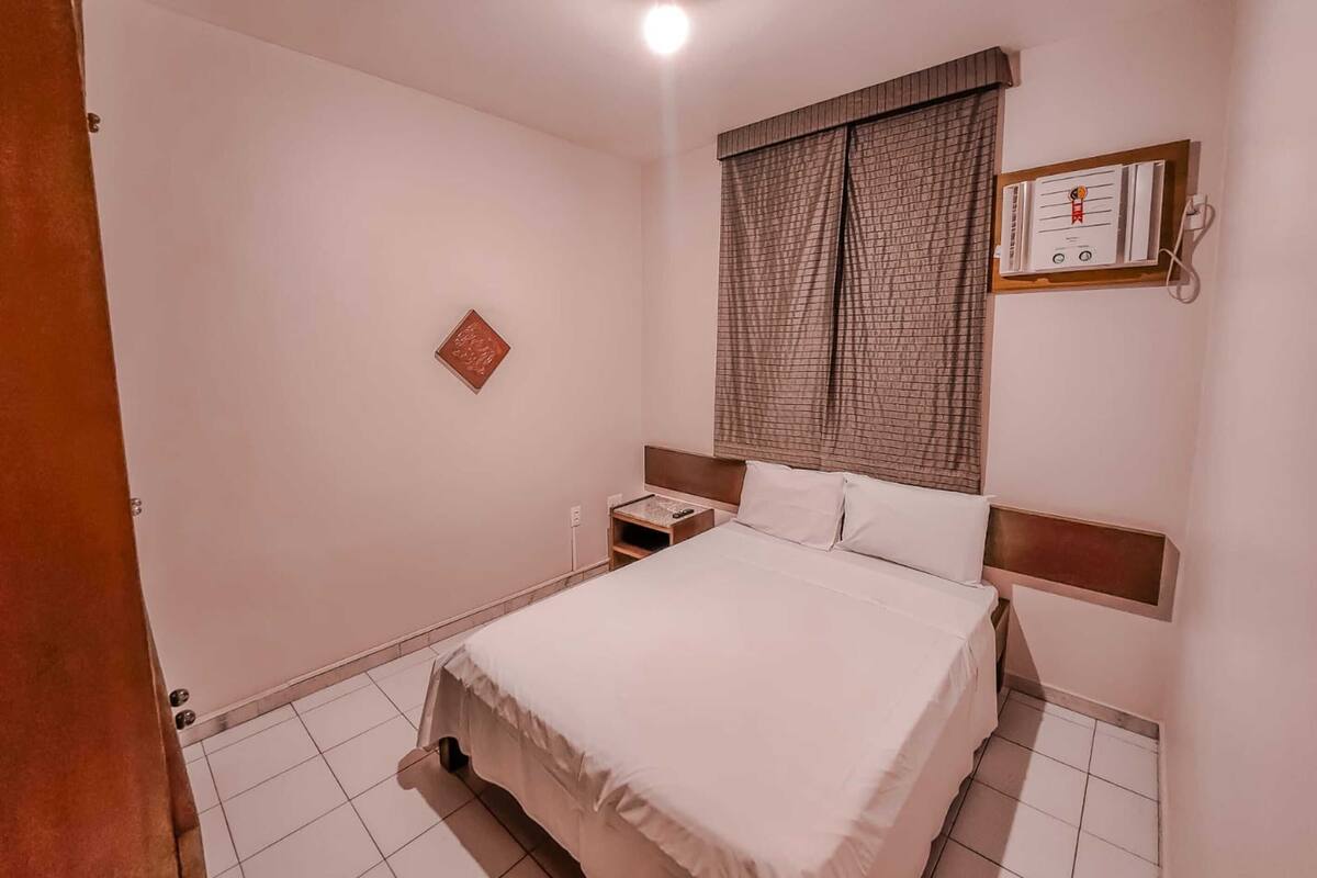 Una camera d'albergo ordinata con un letto, un comodino, una sveglia a parete e una finestra con tende.