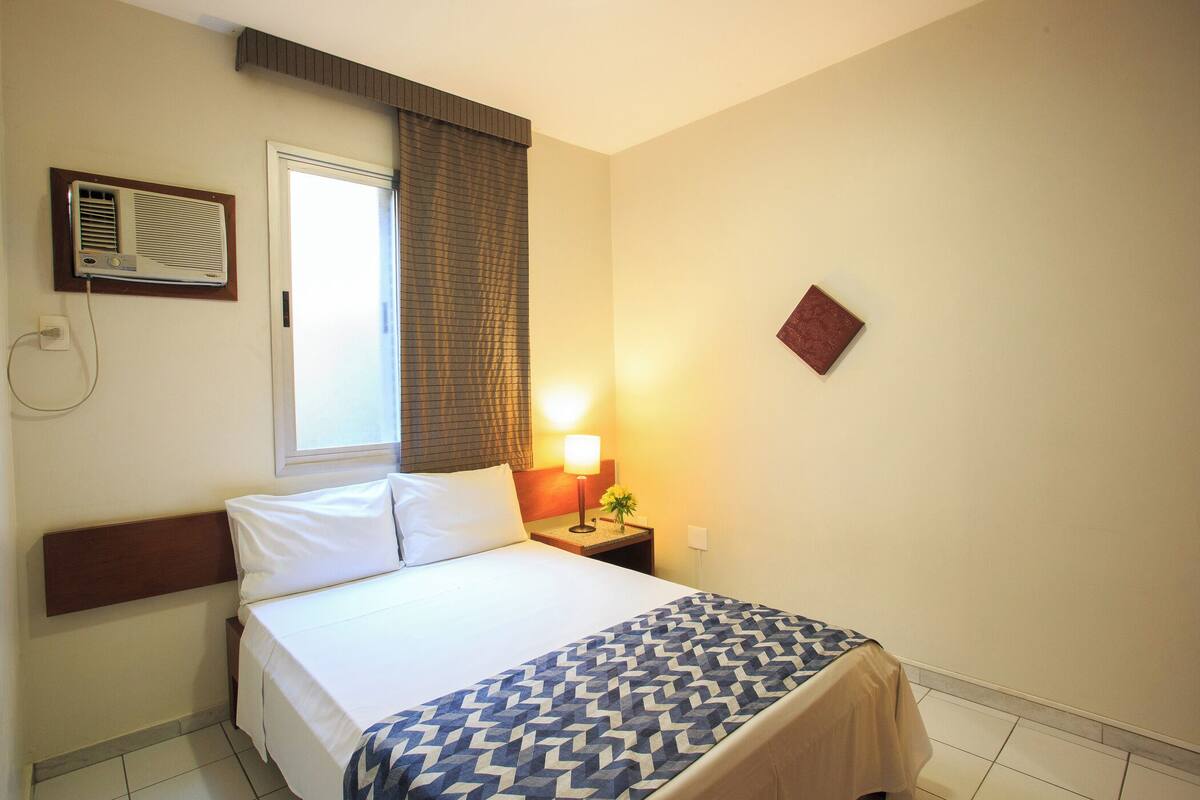 Una camera d'hotel con un letto, un comodino con una lampada, una finestra con tende e un condizionatore d'aria.