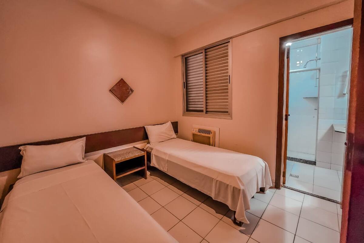 Una camera d'albergo con due letti, un tavolino, un termosifone e una porta scorrevole in vetro che dà sul bagno.