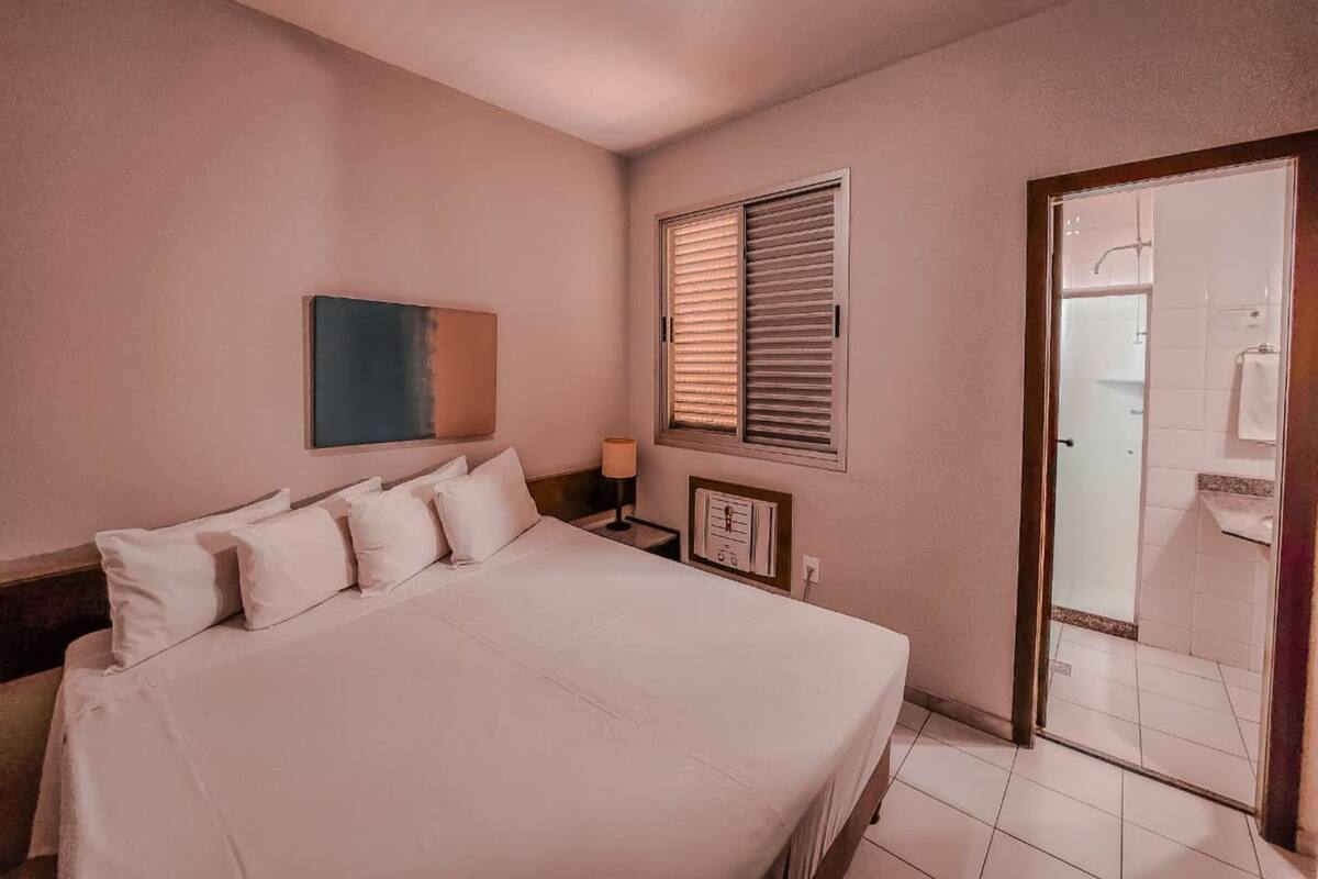 Una camera da letto con un letto grande, un comodino, una lampada a muro, una finestra con persiane e una porta che dà su un bagno.