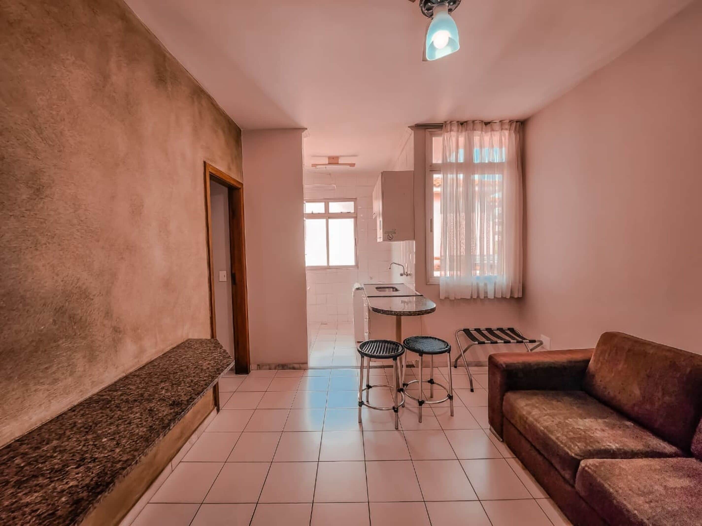 Een woonkamer met een bank, een salontafel met twee stoelen, een kitchenette en een raam met gordijnen.