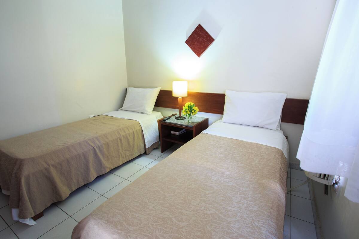 Una camera d'albergo con due letti singoli, un comodino con una lampada e un tavolino con un vaso di fiori.