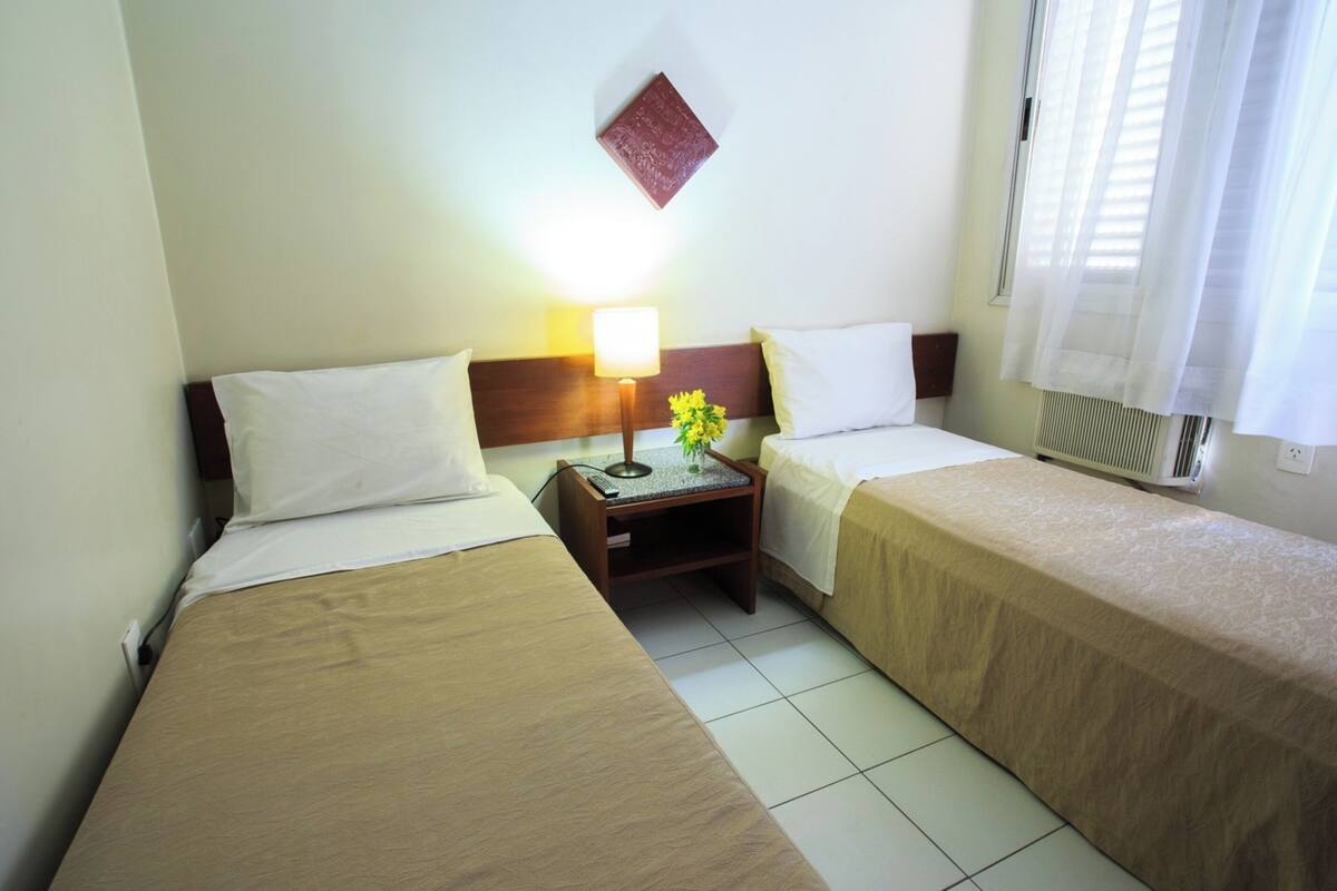 Camera d'albergo con due letti singoli, un comodino con lampada e un piccolo tavolo, e una libreria a parete.