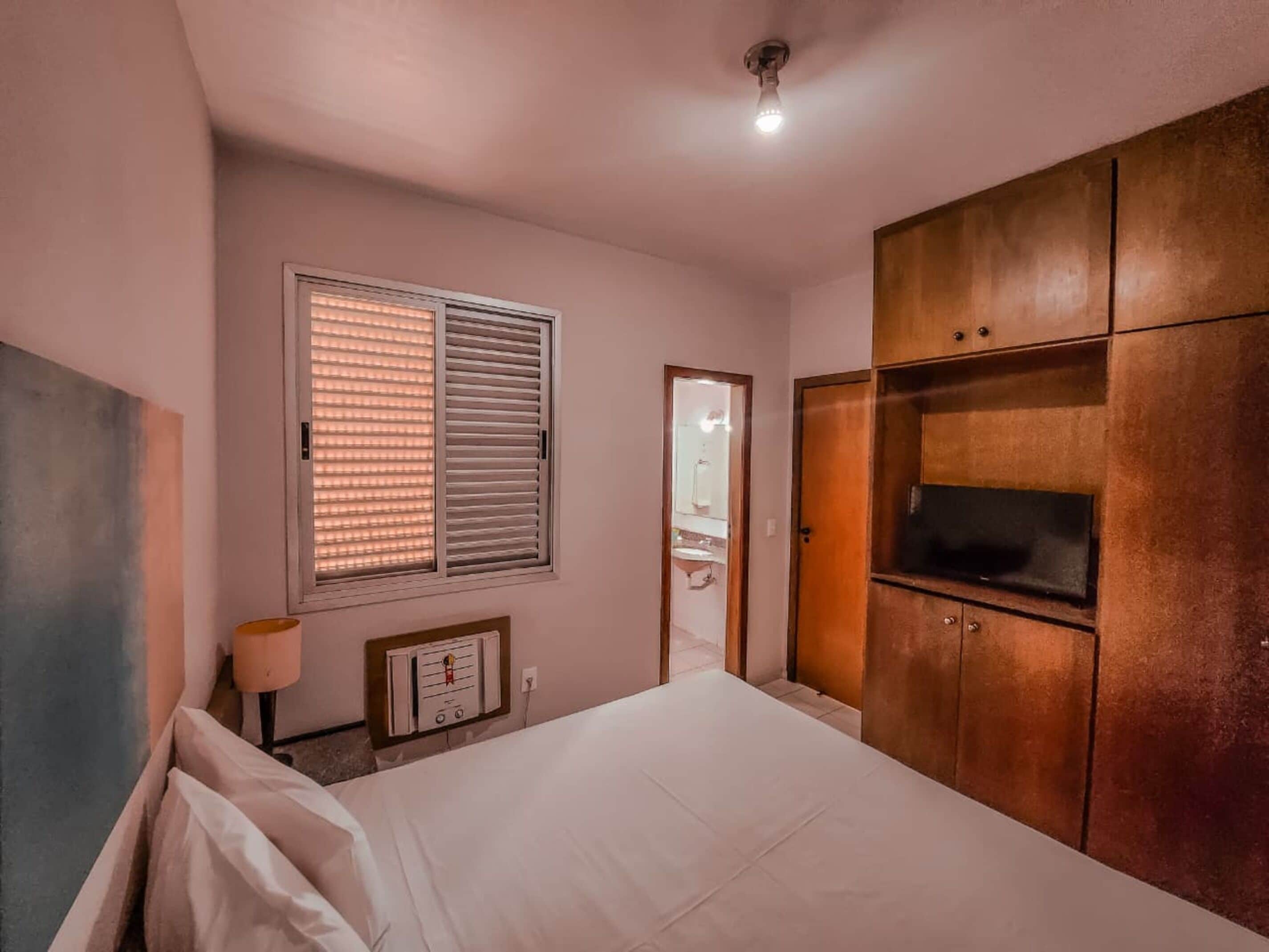 A bedroom with a bed, a TV, a wardrobe, a window with blinds, and a ceiling light.