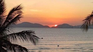 Escena de playa con atardecer, palmeras y sombrillas.