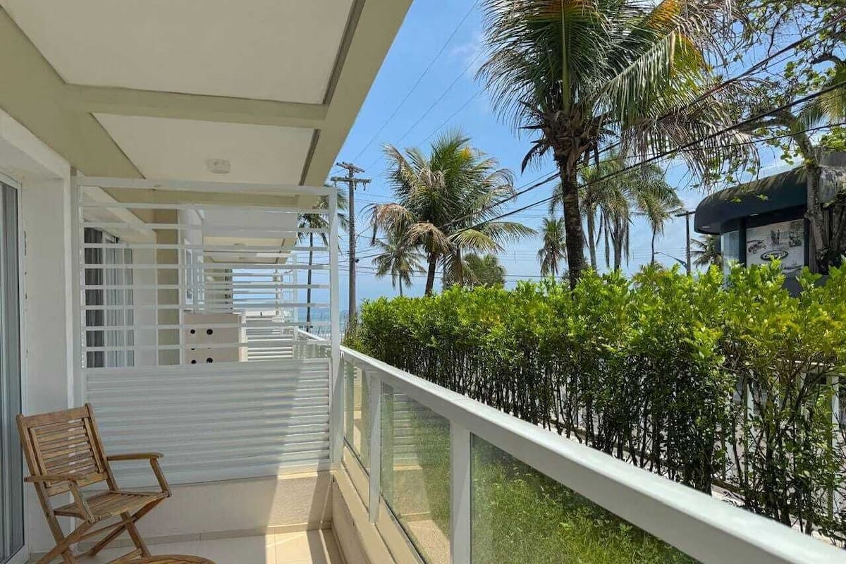A balcony with a white railing, a wooden chair, and a view of palm trees and a building.
