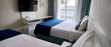 A hotel room with two beds, a flat-screen TV mounted on the wall, a small table with a lamp, and a window with curtains.