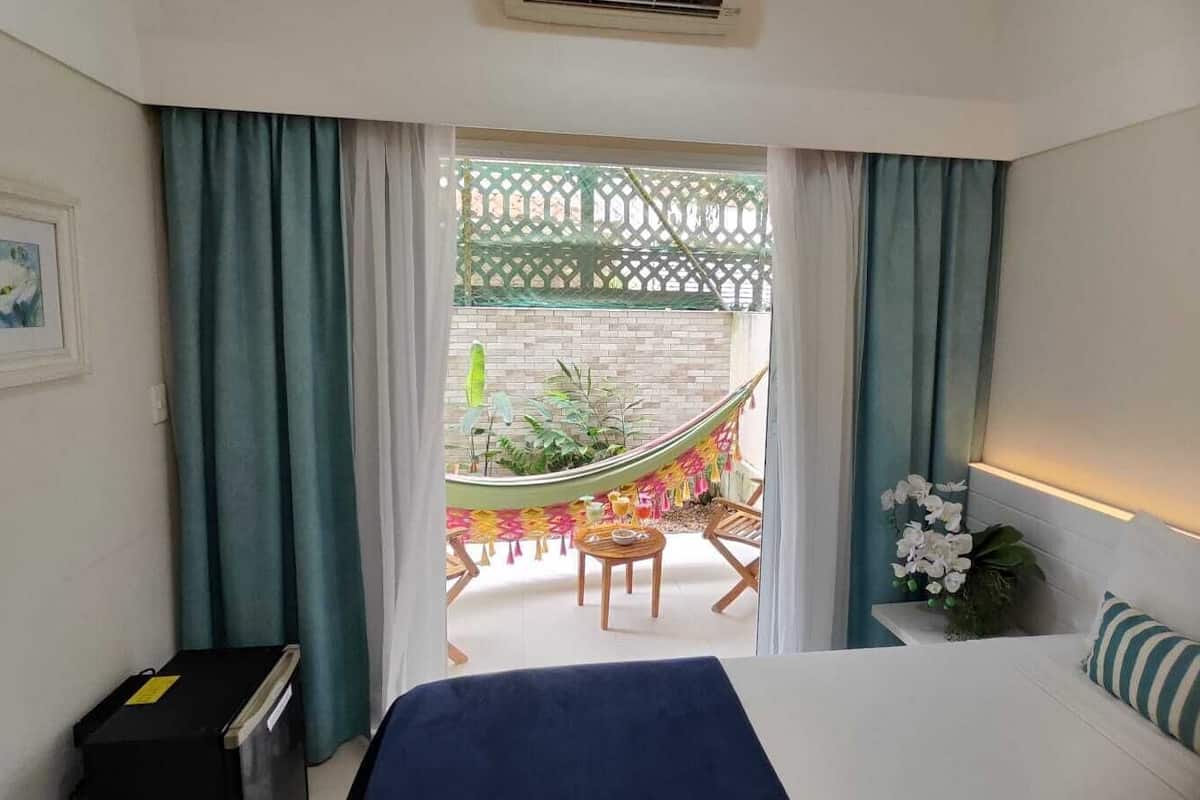A hotel room with a bed, a desk, and a view of an outdoor patio with a hammock and plants.