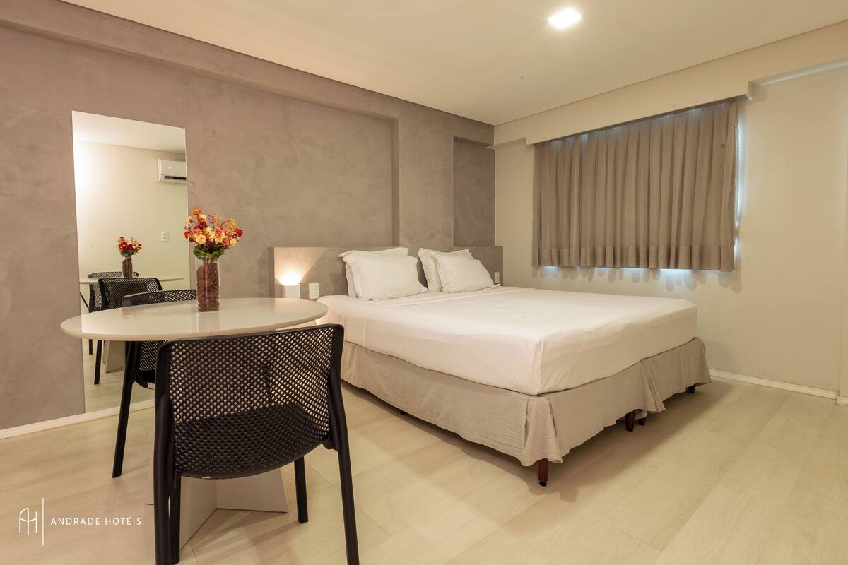 A hotel room with a bed, a dining table with chairs, and a vase of flowers.