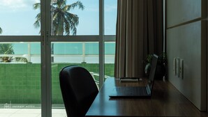 A room with a laptop on a desk, a chair, a large window with a view of palm trees and a green wall. - Rede Andrade Solmar (João Pessoa)