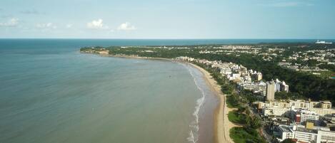 An aerial view of a coastal city with a long sandy beach, dense urban development, and a clear sky.