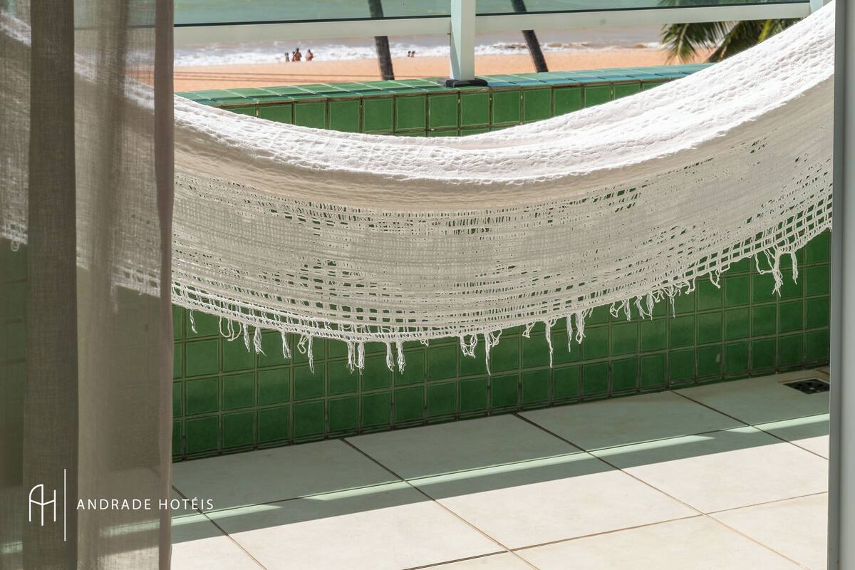 A hammock on a balcony with a view of the beach and palm trees.