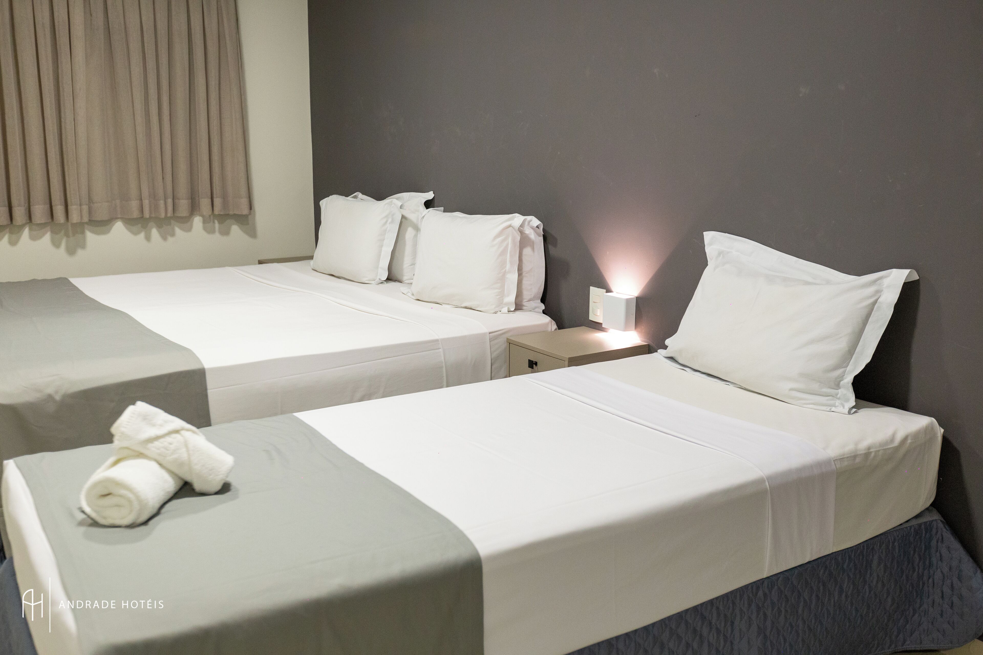 Habitación de hotel con dos camas, cada una con ropa de cama y almohadas blancas, una mesita de noche pequeña con una lámpara y una toalla sobre la primera cama.