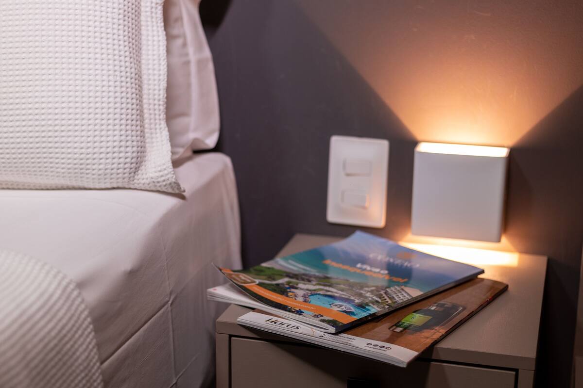 A neatly made bed with a white comforter and a white blanket. A magazine is placed on a bedside table next to a wall-mounted light switch.