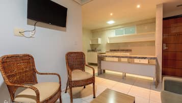 A modern kitchen with a built-in oven, a countertop, and a sink. There are two wicker chairs and a wooden table in the living area.