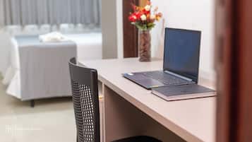 Quarto de hotel com cama, escrivaninha com laptop, cadeira e vaso com flores.