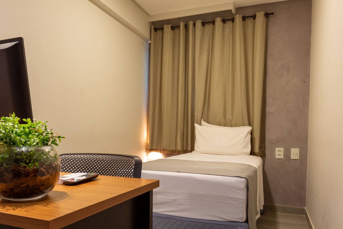 A hotel room with a bed, a TV, a wooden desk, and a potted plant.