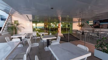 An outdoor seating area with white tables and chairs, a glass wall, and a staircase leading to an upper level.