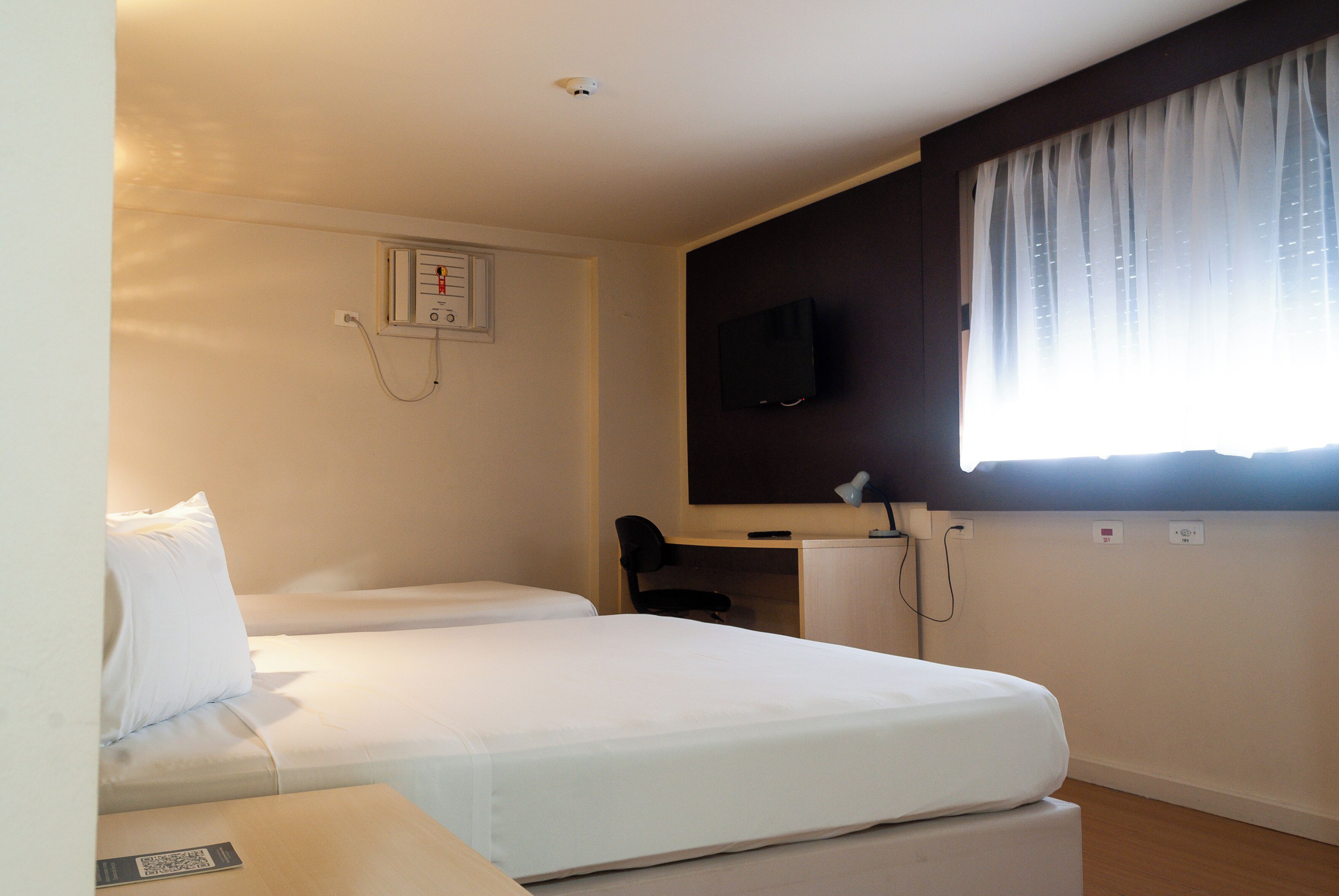 Hotellihuone, jossa on kaksi sänkyä, työpöytä, tuoli, televisio ja ikkuna, jossa on verhot.