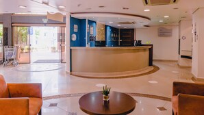 Zona de recepción de hotel con mostrador curvo, escritorio de recepción y una planta pequeña sobre una mesa redonda.