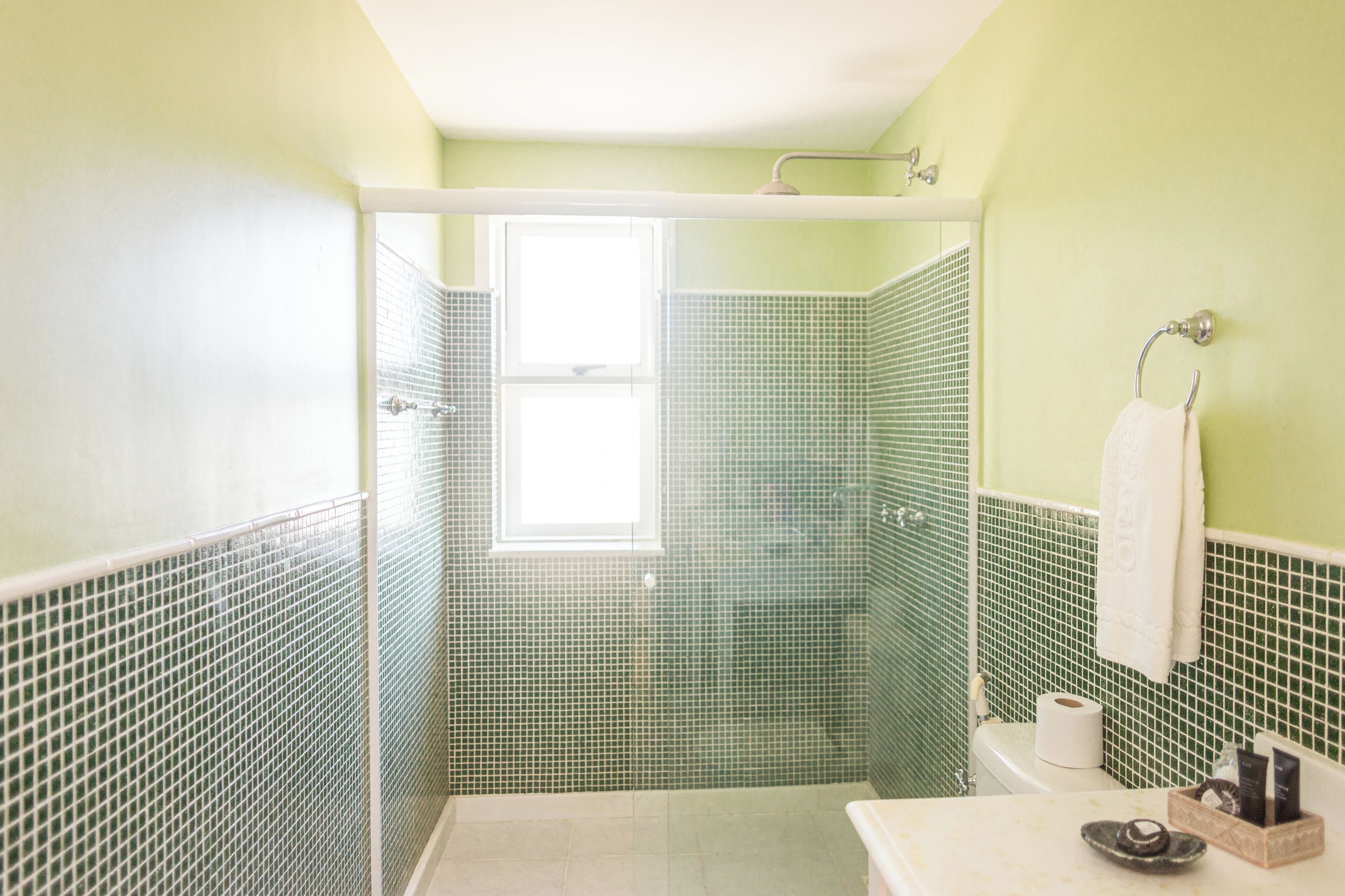 A bathroom with green tiled walls, a shower, a toilet, and a sink with toiletries.