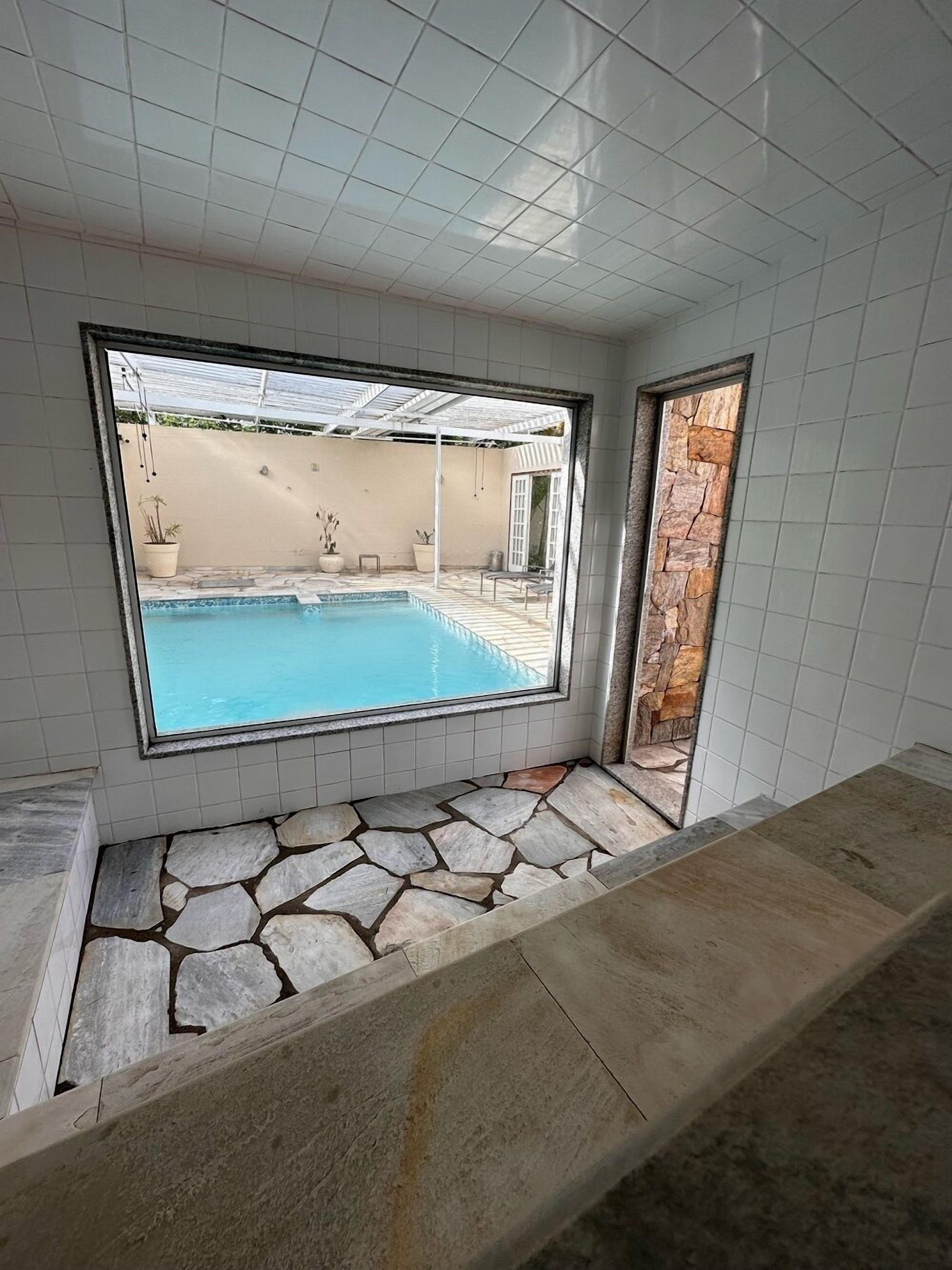 A room with a large window overlooking a swimming pool, stone flooring, and a view of a garden.